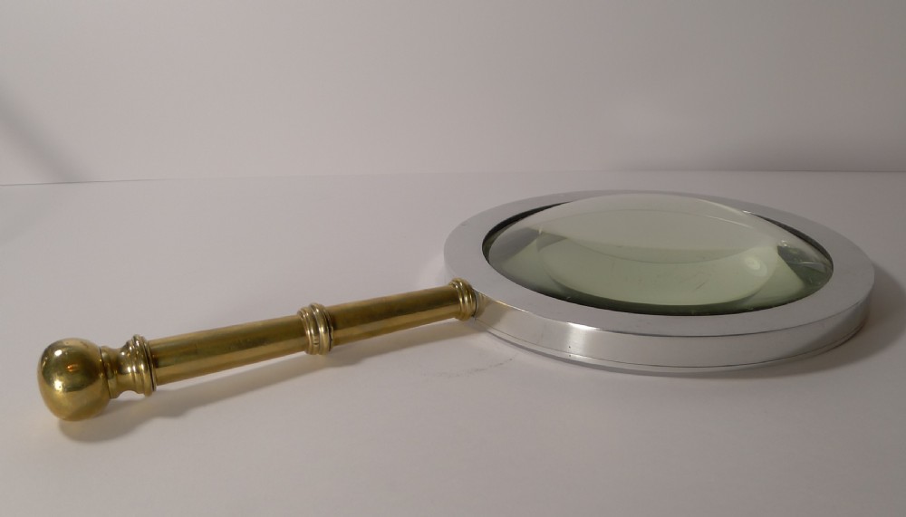 A large round magnifying glass featuring a wide circular silver-toned metal frame around a glass lens and a turned brass handle with a spherical finial. View VII