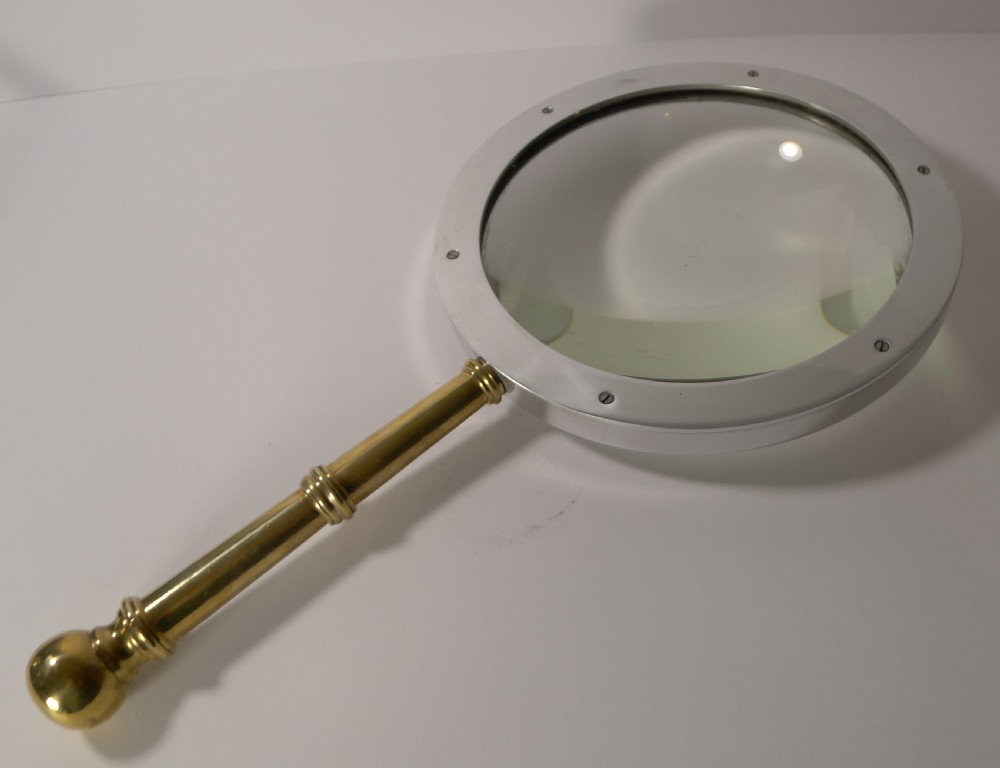 A large round magnifying glass featuring a wide circular silver-toned metal frame around a glass lens and a turned brass handle with a spherical finial. View IX