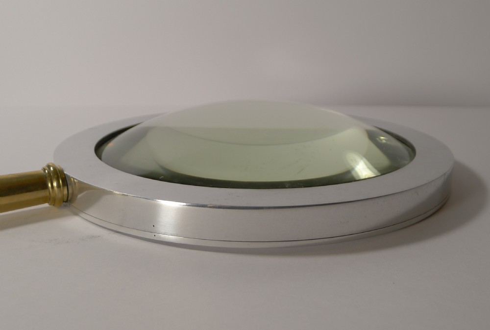 A large round magnifying glass featuring a wide circular silver-toned metal frame around a glass lens and a turned brass handle with a spherical finial. View X