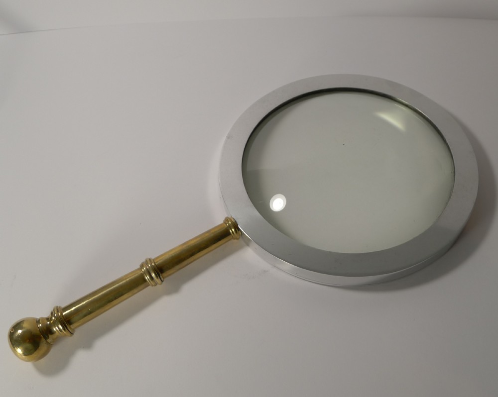 A large round magnifying glass featuring a wide circular silver-toned metal frame around a glass lens and a turned brass handle with a spherical finial. View XII