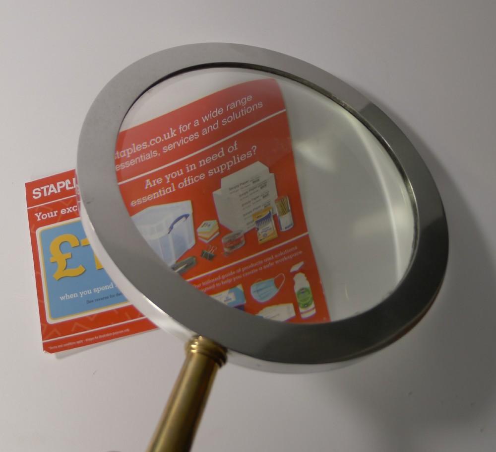 A large round magnifying glass featuring a wide circular silver-toned metal frame around a glass lens and a turned brass handle with a spherical finial. View XIII