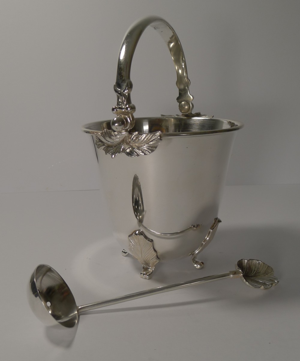 A silver-plated ice bucket featuring an overhead swinging handle, leaf-form feet and rim accents, displayed alongside a matching slotted draining spoon.