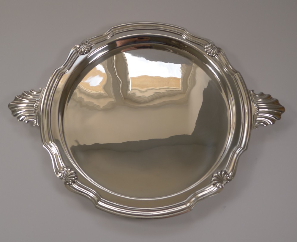 A circular silver-plated serving tray featuring a polished surface, scalloped moulded border, and shell-form handles.