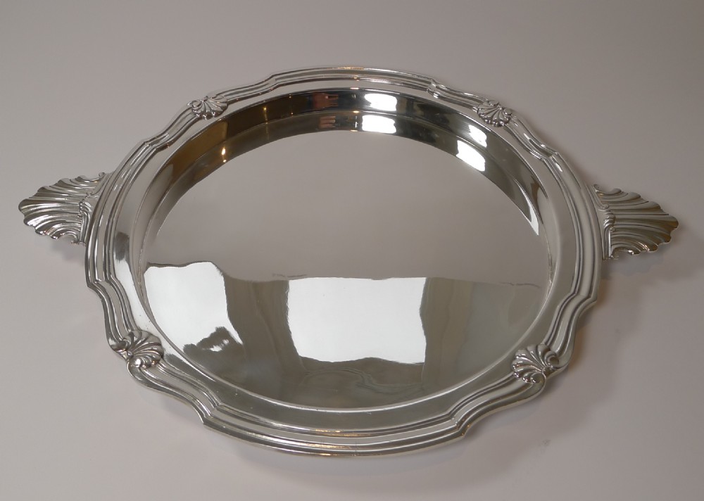 A circular silver-plated serving tray featuring a polished surface, scalloped moulded border, and shell-form handles. View II