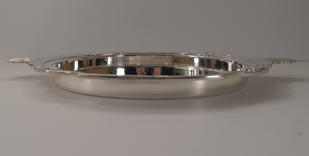 A circular silver-plated serving tray featuring a polished surface, scalloped moulded border, and shell-form handles. View IX