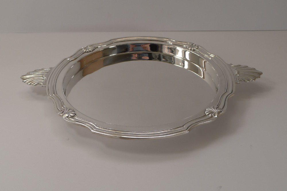 A circular silver-plated serving tray featuring a polished surface, scalloped moulded border, and shell-form handles. View X