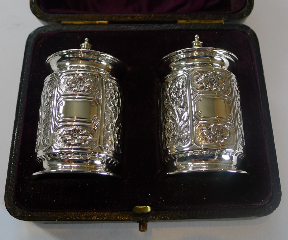 A pair of embossed sterling silver pepper pots with fitted lids displayed inside a dark velvet-lined presentation case.