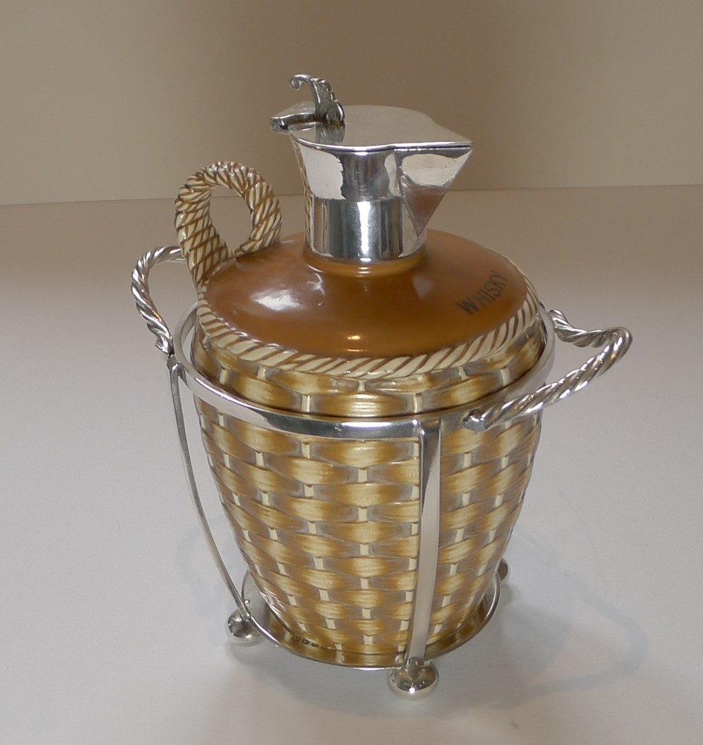An antique Victorian glazed ceramic whisky carafe featuring a basketweave textured lower body, silver-plated spout and lid, and a fitted wire caddy frame.