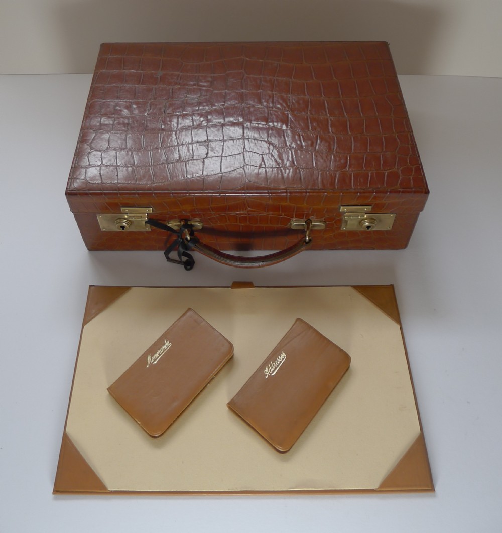 A tan crocodile hide writing case with double brass locks, displayed with a matching leather blotter pad and small notebooks.