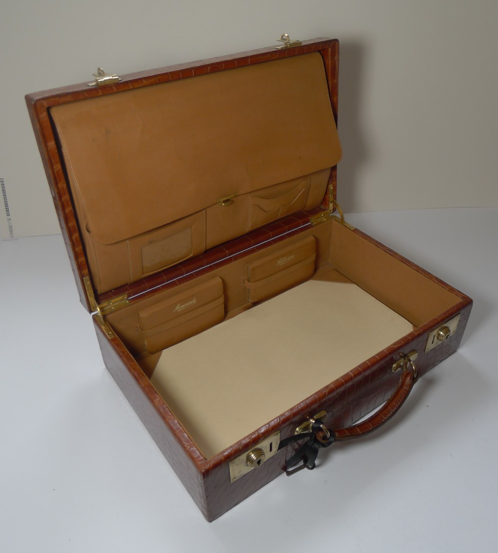 A tan crocodile hide writing case with double brass locks, displayed with a matching leather blotter pad and small notebooks. View VII