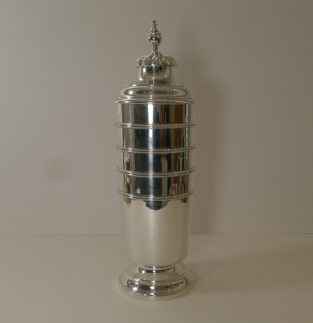 A tall, polished sterling silver cocktail shaker featuring horizontal ribbed banding, a stepped base, and a decorative finial cap.