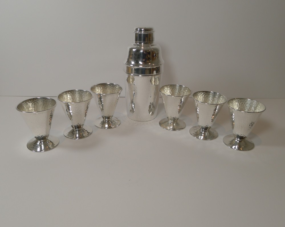 An Art Deco silver-plated cocktail shaker set with a hammered texture, featuring a central shaker flanked by six matching flared goblets.