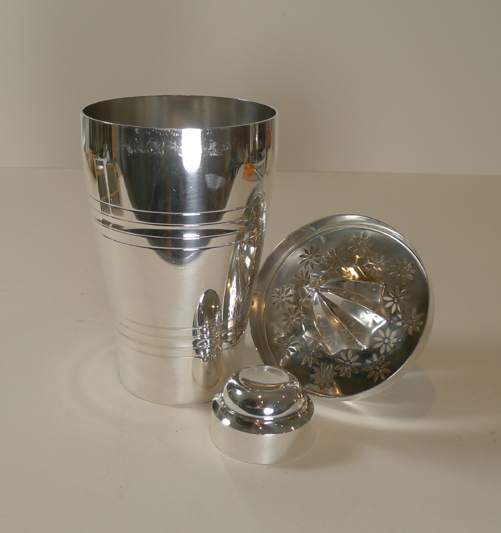A silver-plated Art Deco cocktail shaker displayed alongside its detached floral-embossed lemon reamer lid and small cap.