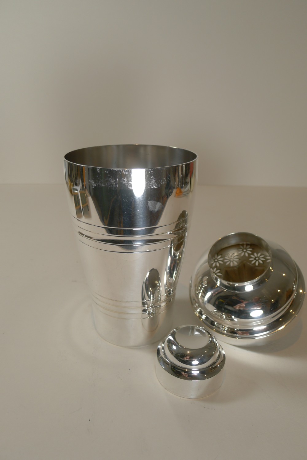 A silver-plated Art Deco cocktail shaker displayed alongside its detached floral-embossed lemon reamer lid and small cap. View IX