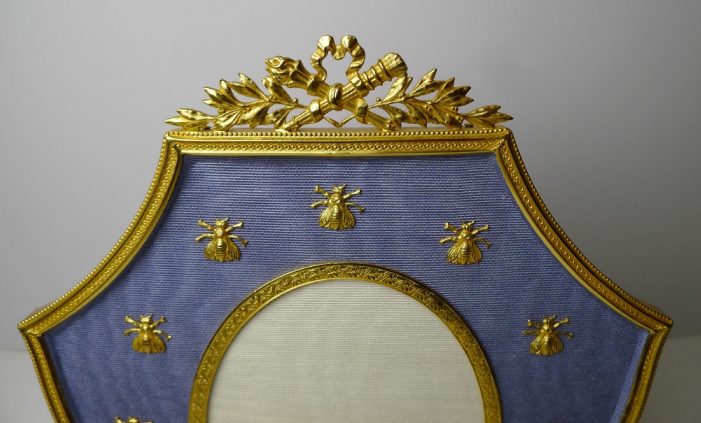 A shield-shaped gilded bronze picture frame with a floral crest top, lined in blue textured fabric decorated with small gold bee motifs surrounding an oval portrait aperture. View IV