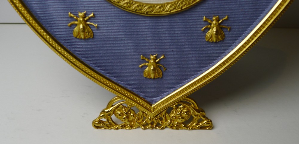 A shield-shaped gilded bronze picture frame with a floral crest top, lined in blue textured fabric decorated with small gold bee motifs surrounding an oval portrait aperture. View VI