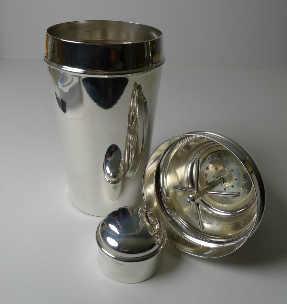 An Art Deco silver-plated cocktail shaker presented disassembled, showing the main body, cap, and strainer lid with an integrated internal ice breaker mechanism.