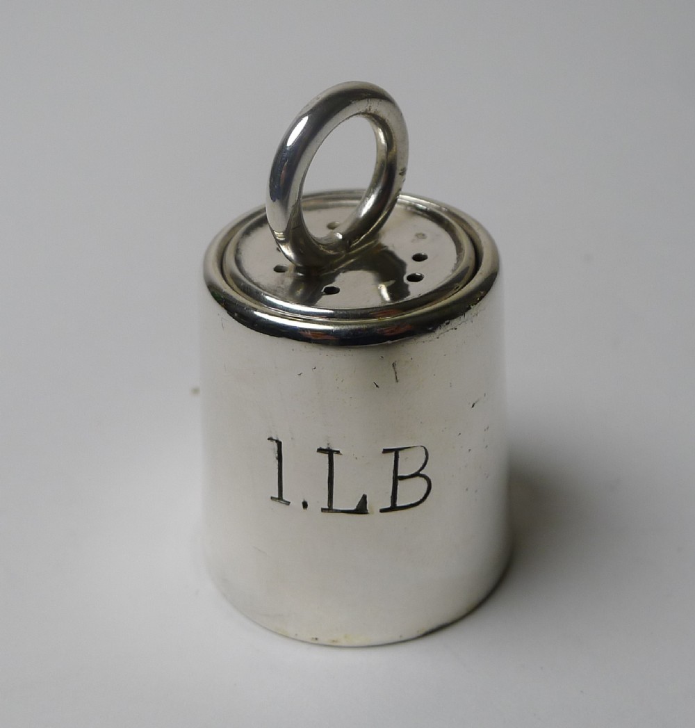 A silver-plated novelty pepper pot designed in the shape of a one-pound trade weight with a top ring handle and engraved text.