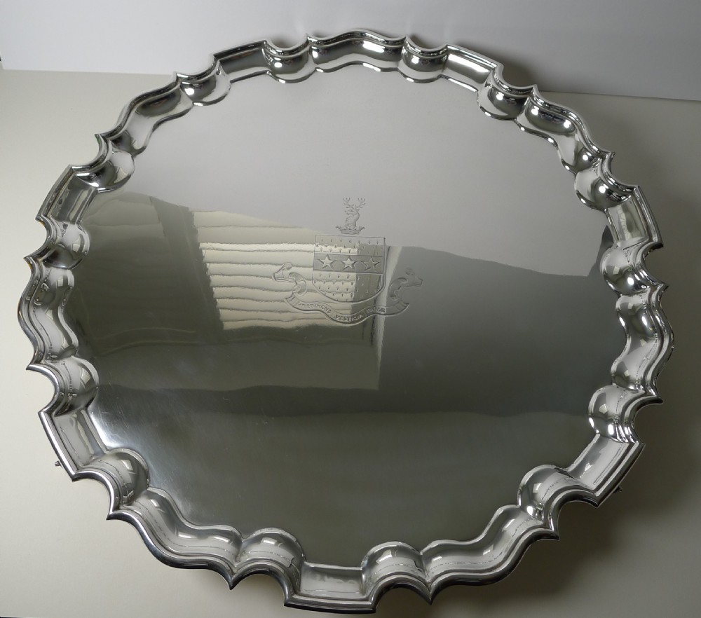 A circular silver-plated salver featuring a scalloped piecrust rim and an engraved armorial family crest in the centre.