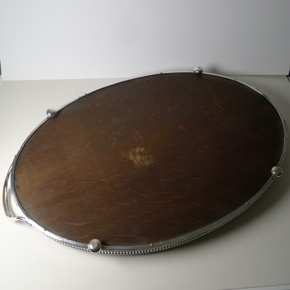 An Edwardian oval dark oak tray featuring a silver pierced gallery border, twin side handles, small bun feet, and a central oval silver medallion. View VI
