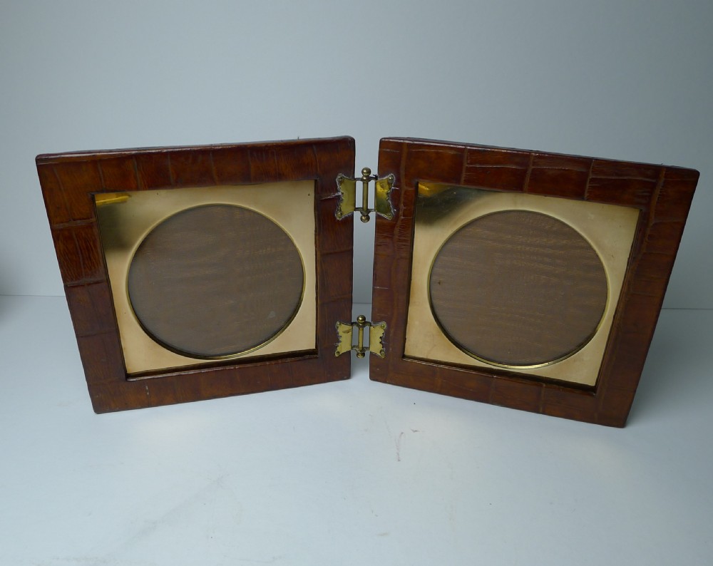 A double folding picture frame bound in brown crocodile-embossed leather featuring circular brass inner apertures and brass hinges.