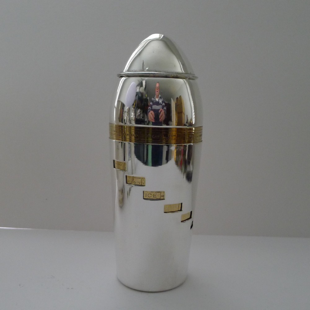 A bullet-shaped silver-plated cocktail shaker featuring a rotating brass band and cut-out windows displaying recipe ingredients.