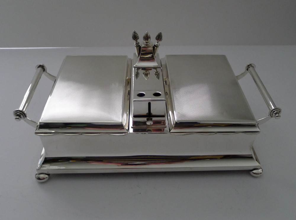 An oblong silver smoking compendium by Mappin & Webb featuring twin hinged lids, central lighter unit with finials, side handles, and a ball-footed base. View XVI
