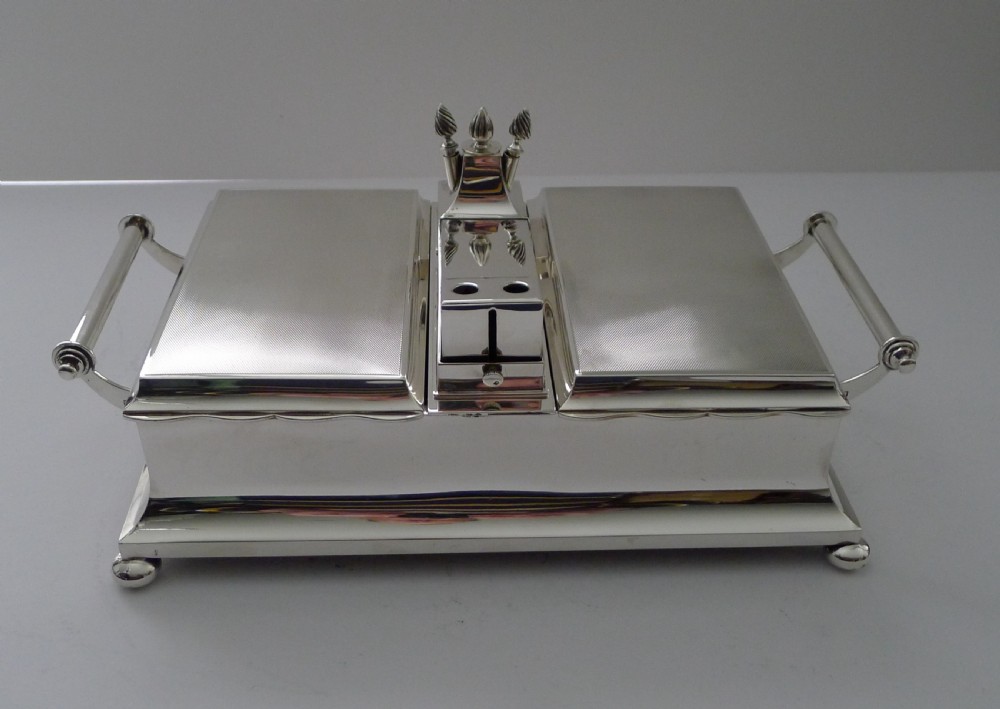 An oblong silver smoking compendium by Mappin & Webb featuring twin hinged lids, central lighter unit with finials, side handles, and a ball-footed base. View XVIII