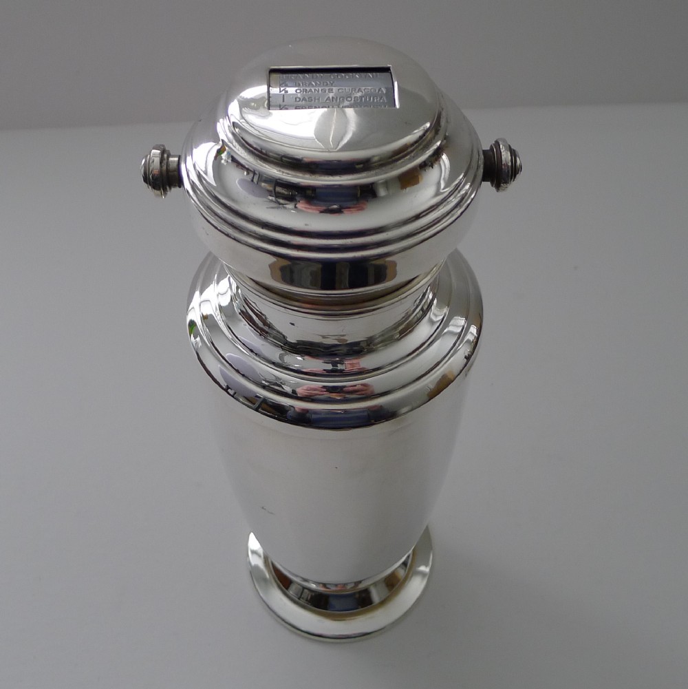 A silver-plated Art Deco cocktail shaker featuring a rotating top cap with rectangular windows displaying drink recipes.