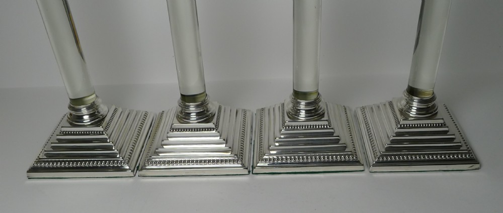 A set of four Victorian silver-plated candlesticks featuring clear glass column shafts, cast Corinthian capitals, and square stepped bases. View X