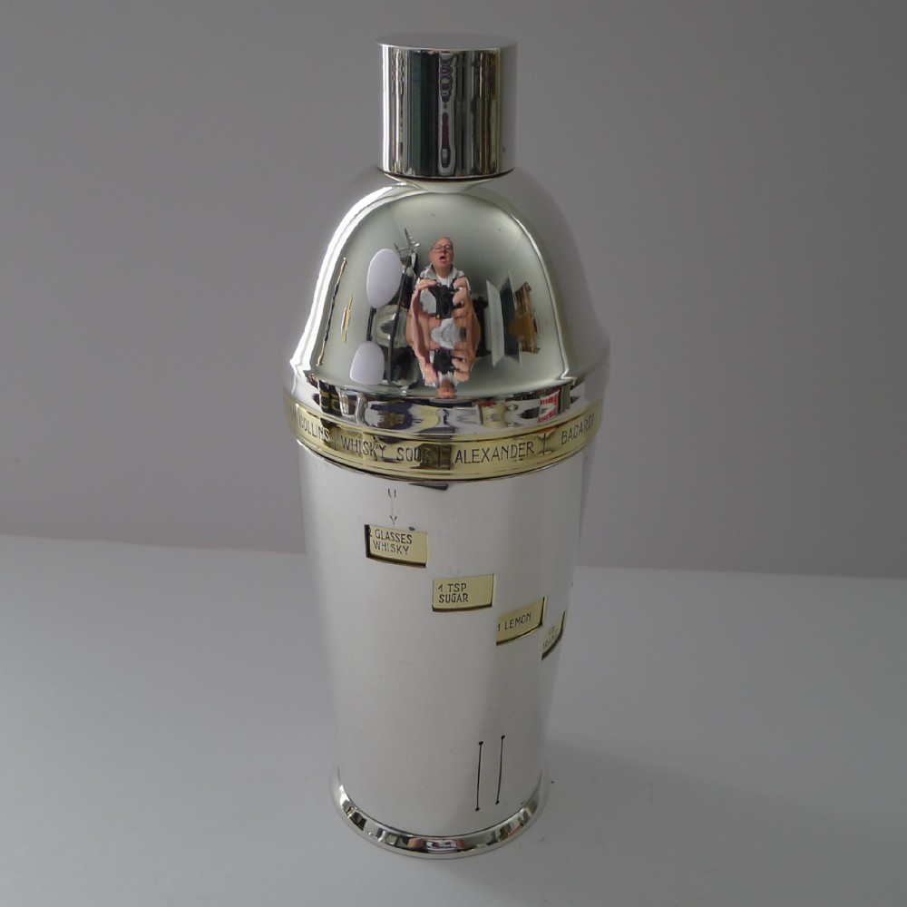A cylindrical silver-plated Art Deco cocktail shaker featuring a rotating brass recipe sleeve with windowed ingredient listings.