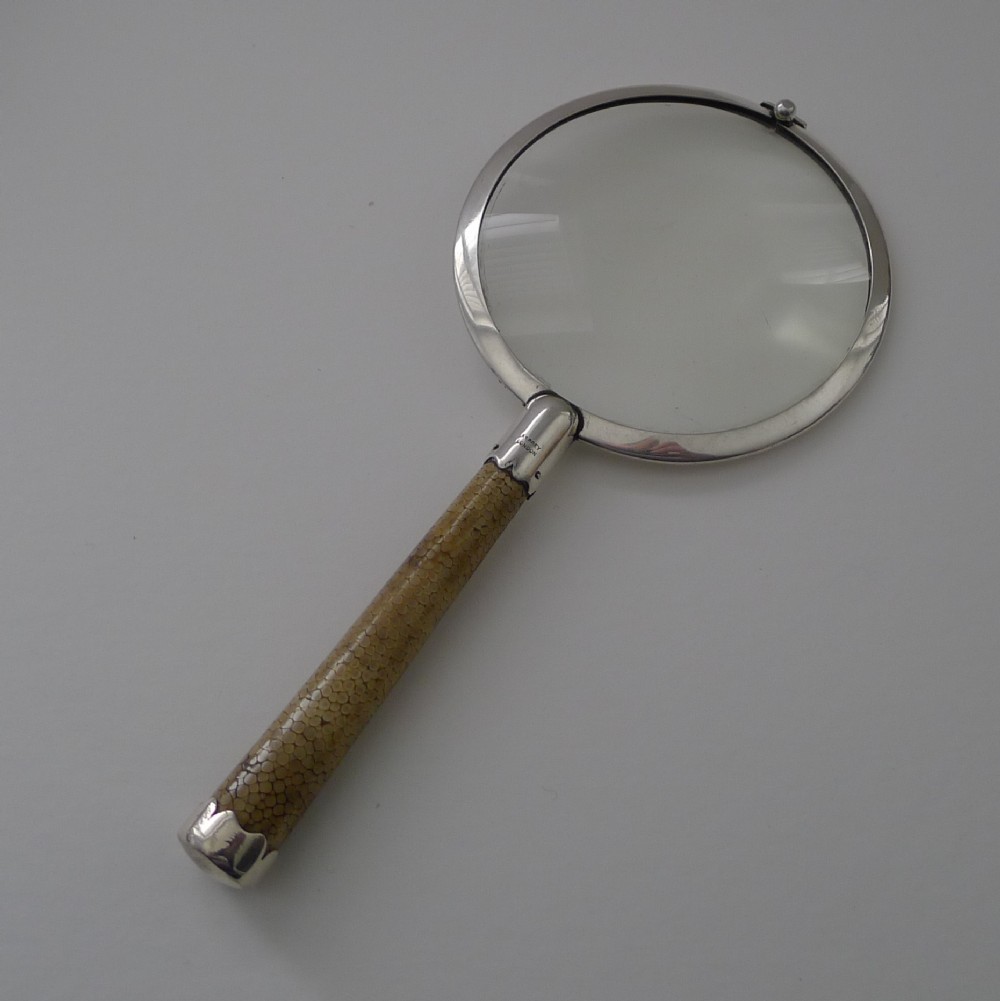 An Art Deco handheld magnifying glass featuring a circular glass lens in a sterling silver frame attached to a tan shagreen handle.