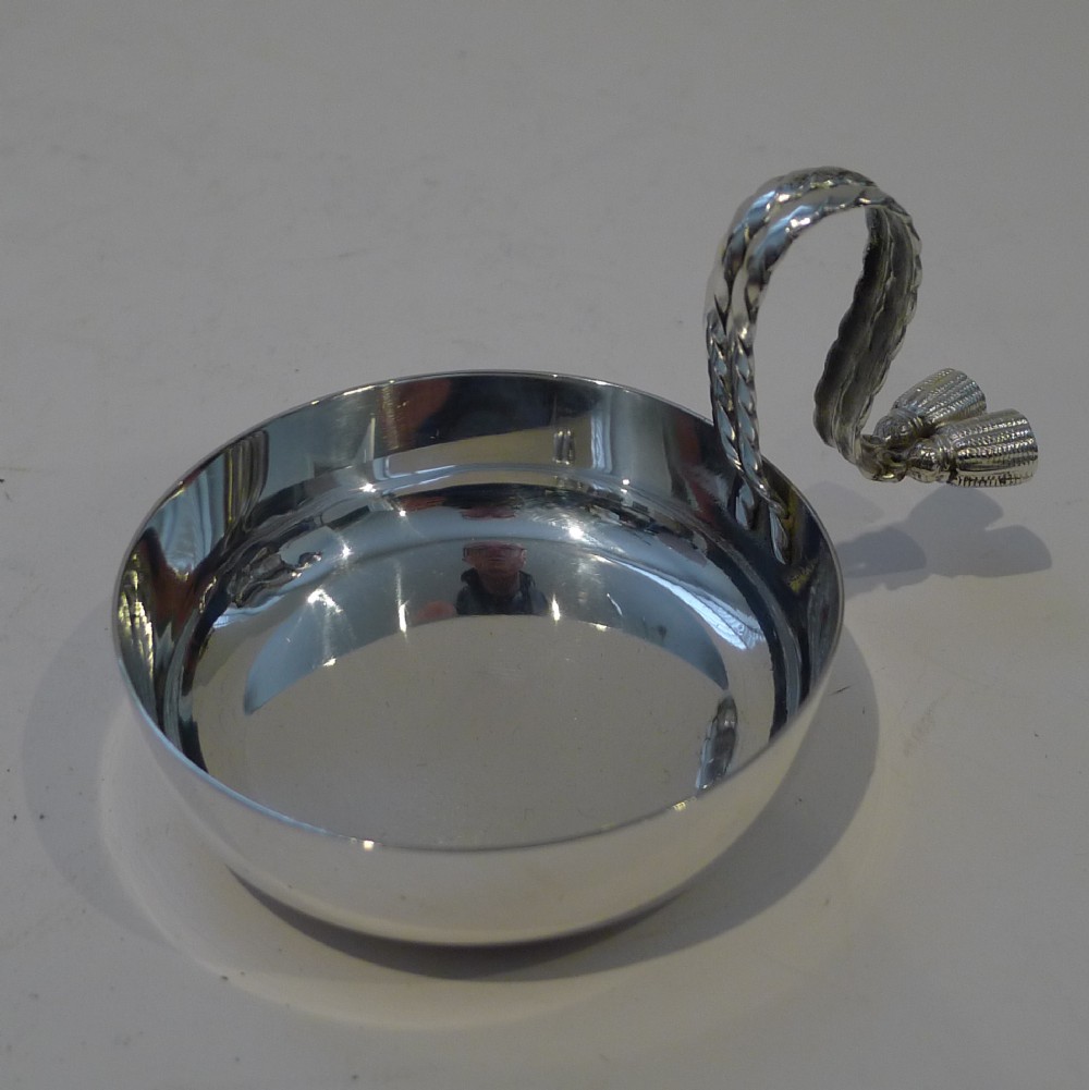 A circular silver-plated shallow dish with a polished reflective finish, featuring an arched twisted handle terminating in decorative double tassels.
