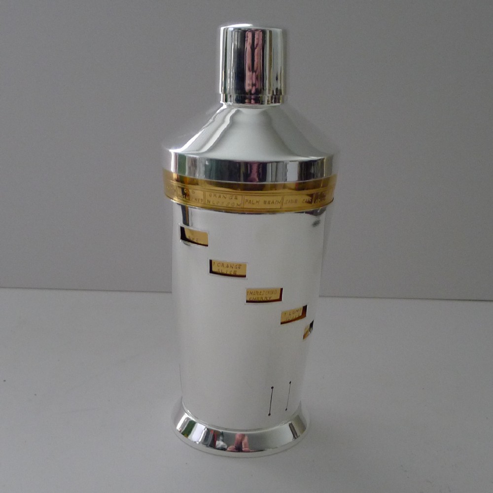 A silver-plated Art Deco cocktail shaker featuring a gilded recipe band and rectangular window slots displaying drink ingredients.