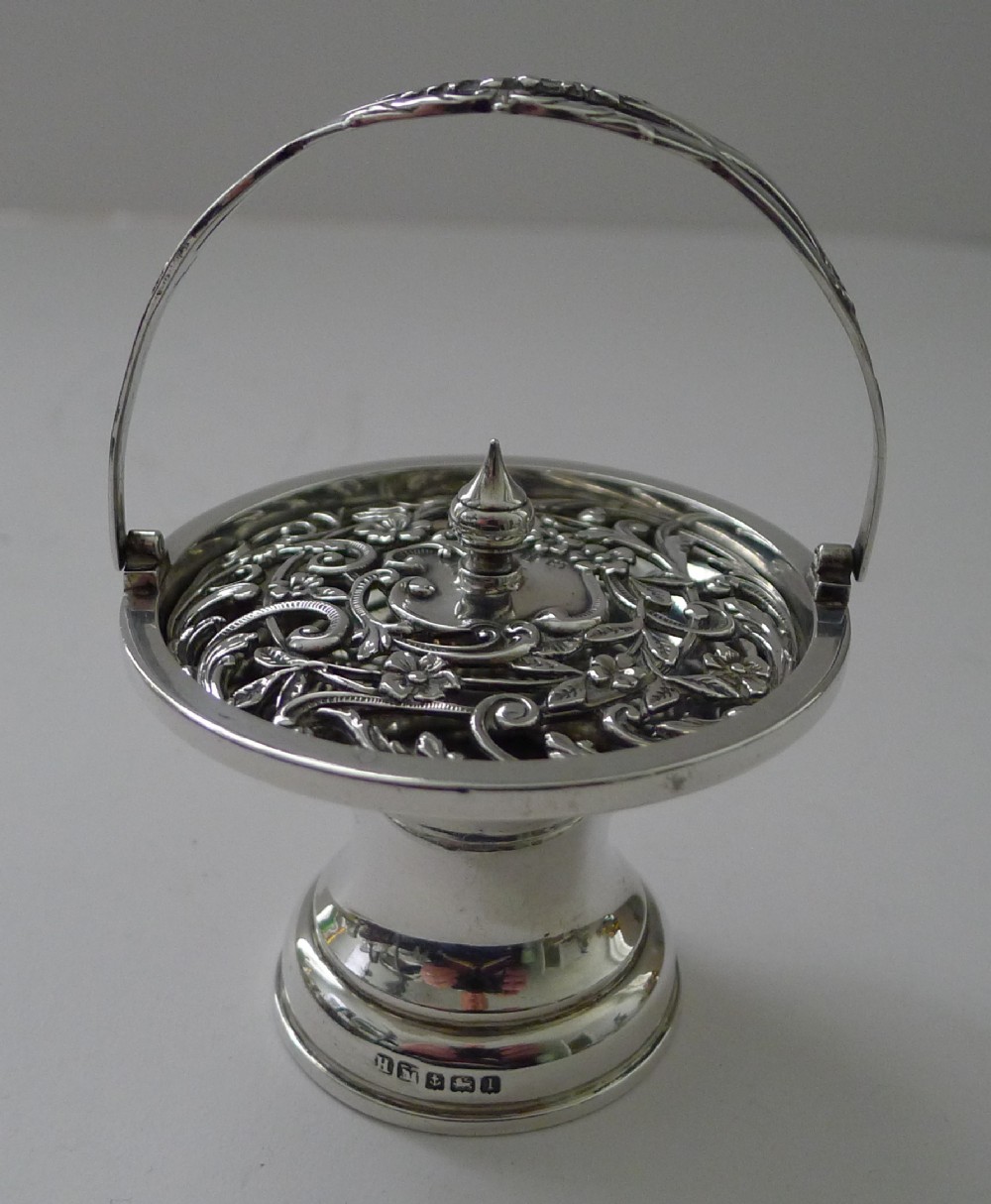 An Edwardian sterling silver pedestal pot pourri basket featuring an overhead swing handle and a pierced floral scrollwork lid topped with a pointed finial.