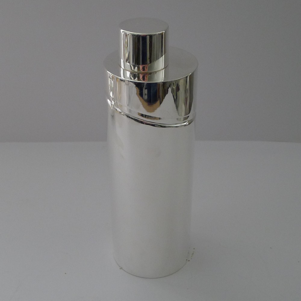 A sleek, highly polished cylindrical silver-plated cocktail shaker with an angled upper seam and fitted cylindrical cap.