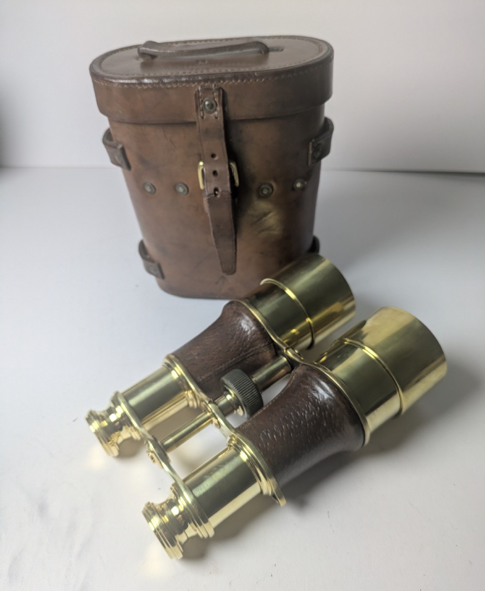 A pair of antique brass field binoculars with textured brown leather grips alongside a fitted brown leather carrying case with straps. View II