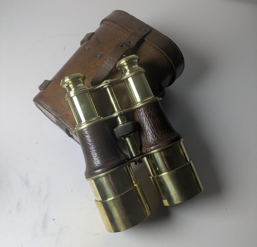 A pair of antique brass field binoculars with textured brown leather grips alongside a fitted brown leather carrying case with straps. View III