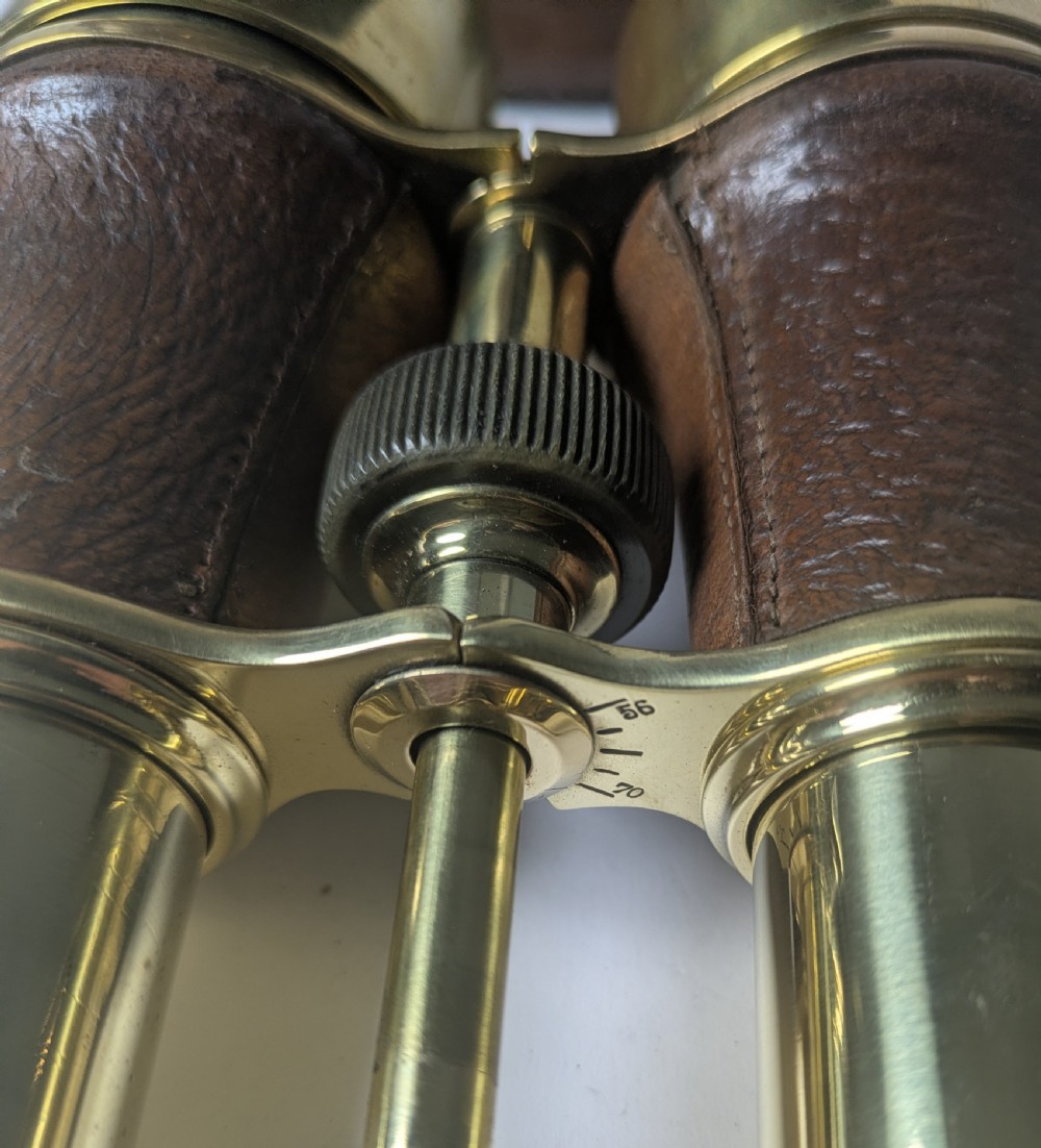 A pair of antique brass field binoculars with textured brown leather grips alongside a fitted brown leather carrying case with straps. View VII
