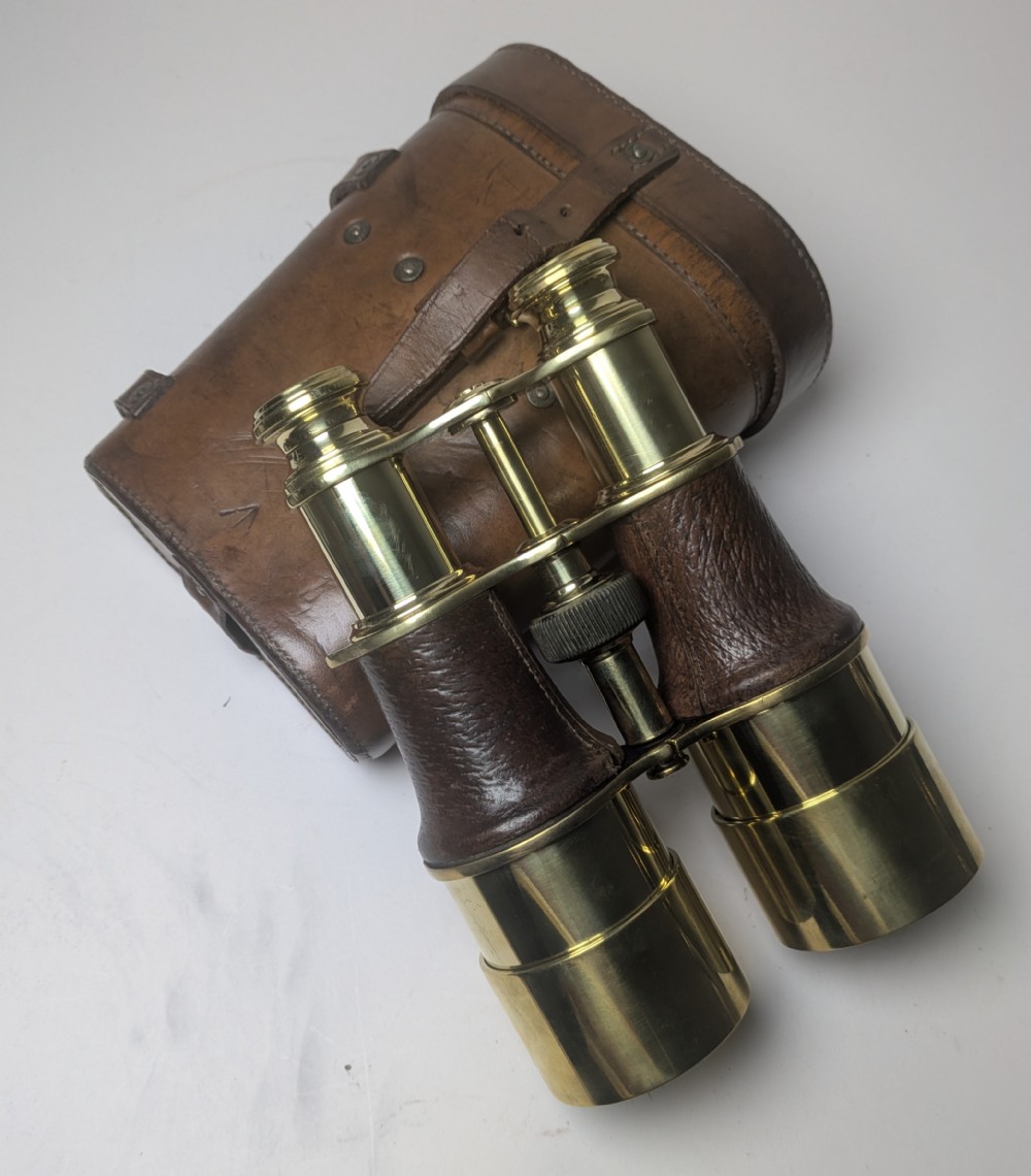 A pair of antique brass field binoculars with textured brown leather grips alongside a fitted brown leather carrying case with straps.