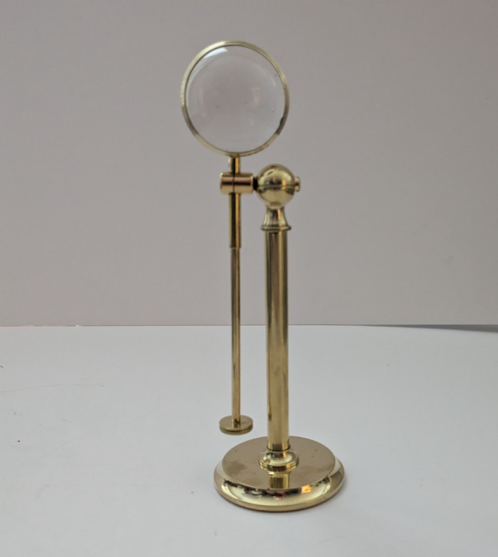 An Edwardian brass standing magnifying glass featuring a circular base, a vertical stem, and an adjustable horizontal arm holding a thick bullseye glass lens. View VII