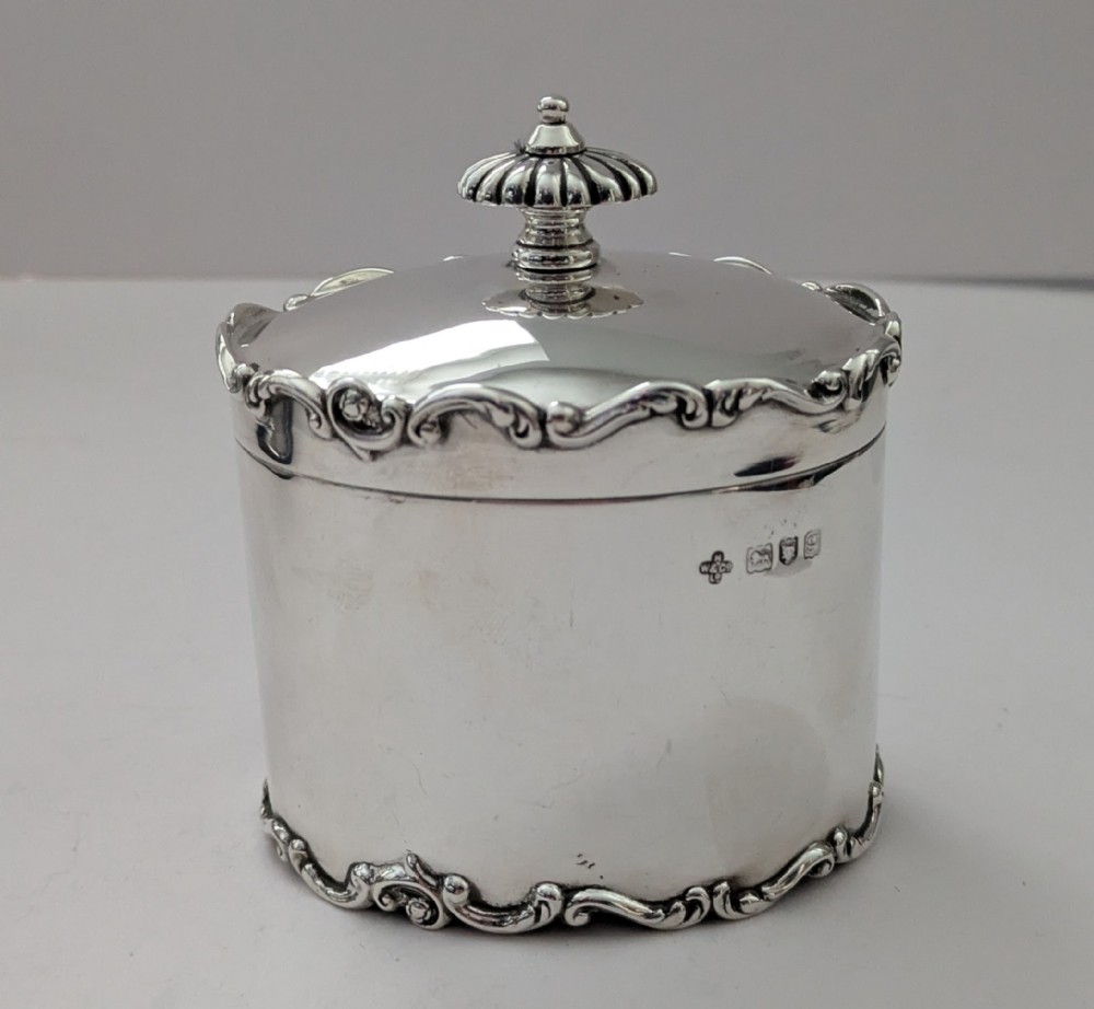 A cylindrical sterling silver tea caddy featuring a scrolled rim border, a fluted finial lid, and stamped London hallmarks on the side.
