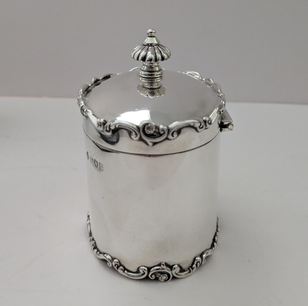 A cylindrical sterling silver tea caddy featuring a scrolled rim border, a fluted finial lid, and stamped London hallmarks on the side. View II