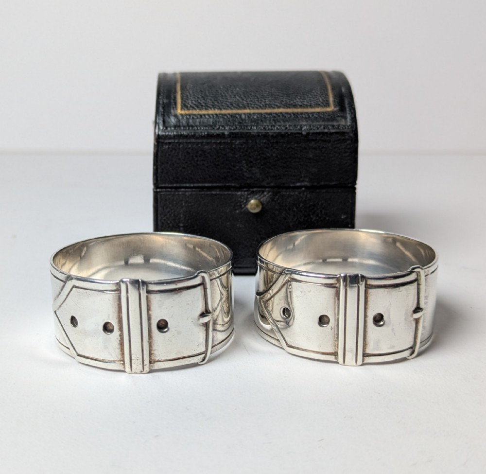 A pair of late Victorian sterling silver napkin rings crafted in the form of buckled belts, shown in front of a black leather presentation case.