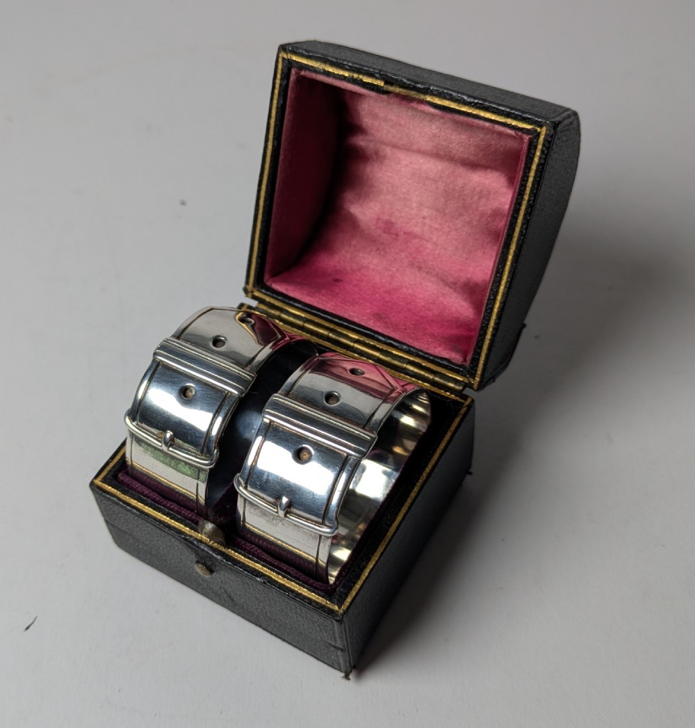 A pair of late Victorian sterling silver napkin rings crafted in the form of buckled belts, shown in front of a black leather presentation case. View IX