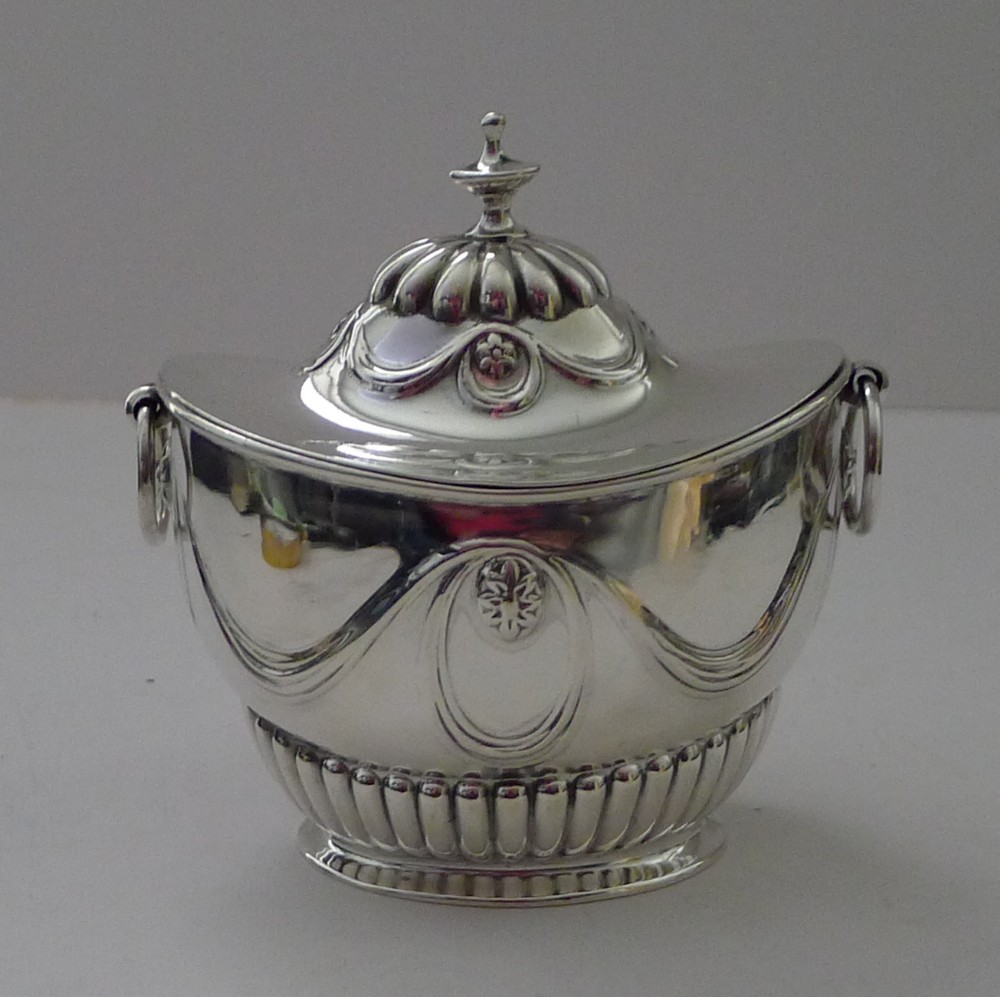An antique Edwardian sterling silver tea caddy featuring lower fluted detailing, embossed swag motifs, drop ring handles, and a finial lid.