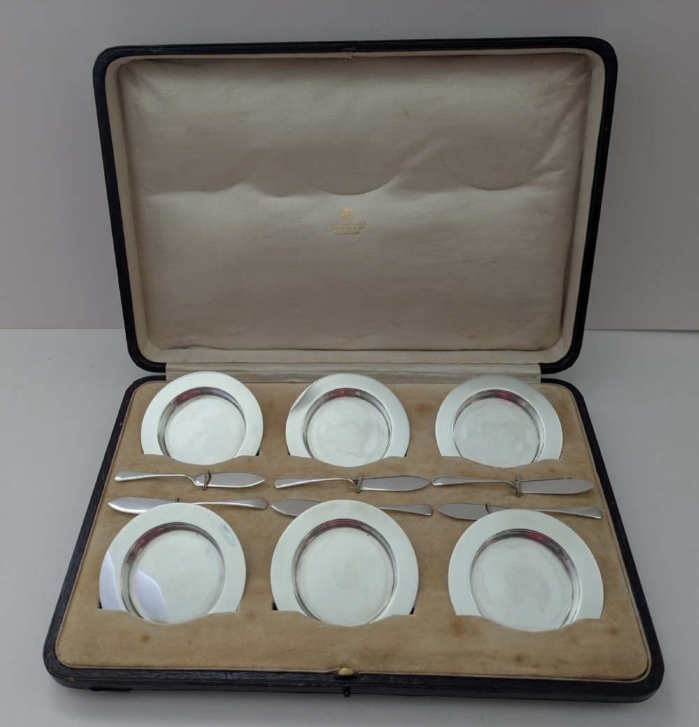 A boxed set of six circular sterling silver butter dishes paired with six matching butter knives inside a silk-lined presentation case.