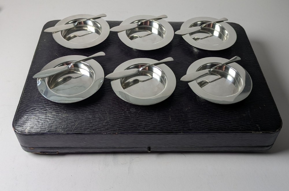 A boxed set of six circular sterling silver butter dishes paired with six matching butter knives inside a silk-lined presentation case. View II