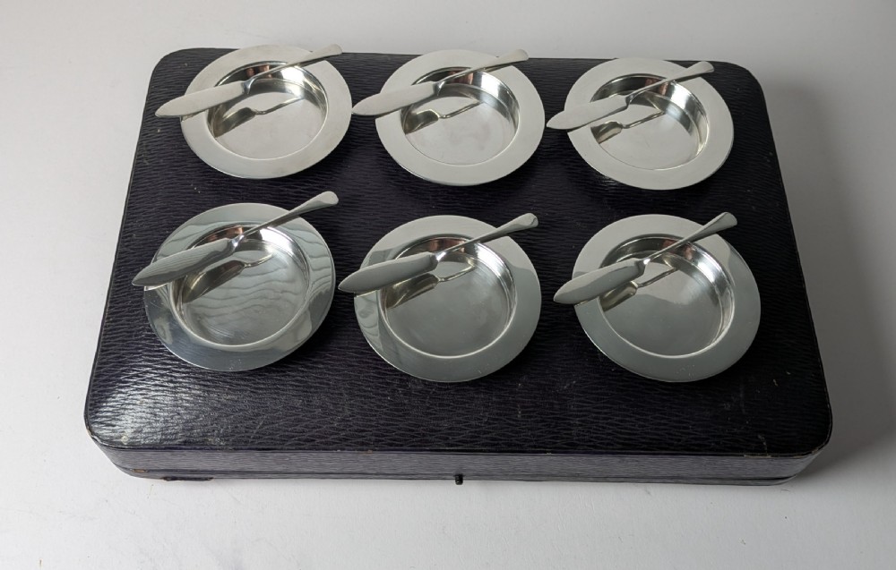 A boxed set of six circular sterling silver butter dishes paired with six matching butter knives inside a silk-lined presentation case. View III