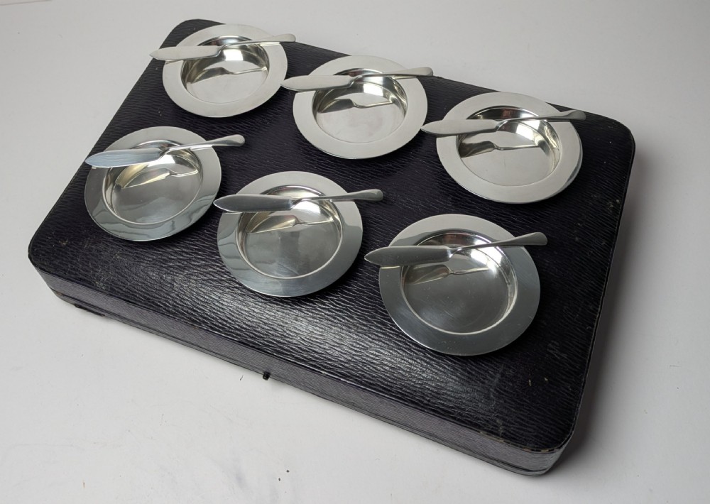 A boxed set of six circular sterling silver butter dishes paired with six matching butter knives inside a silk-lined presentation case. View IV